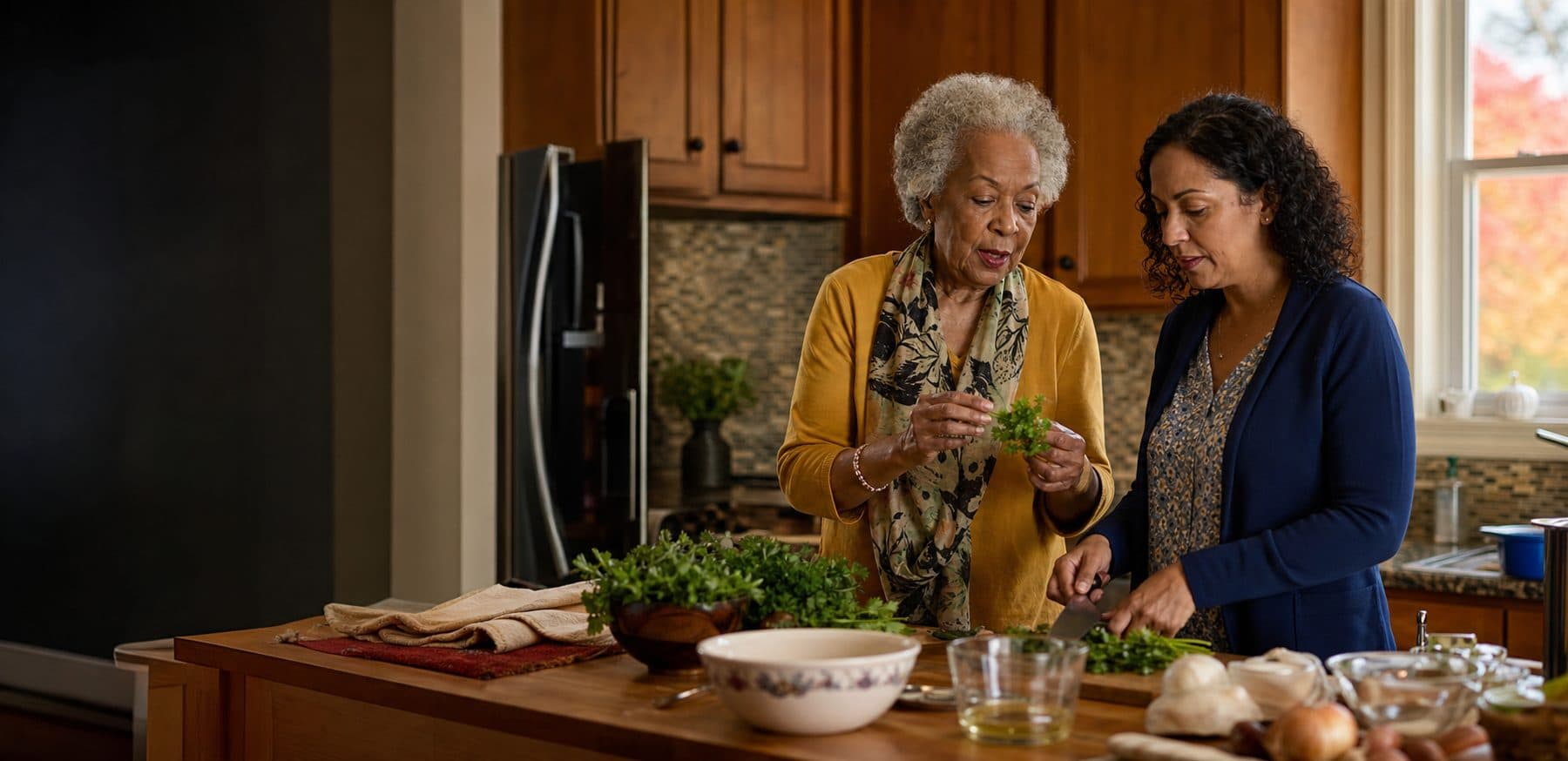 An older woman leads meal preparation in her own kitchen while a home-care aide chops herbs beside her